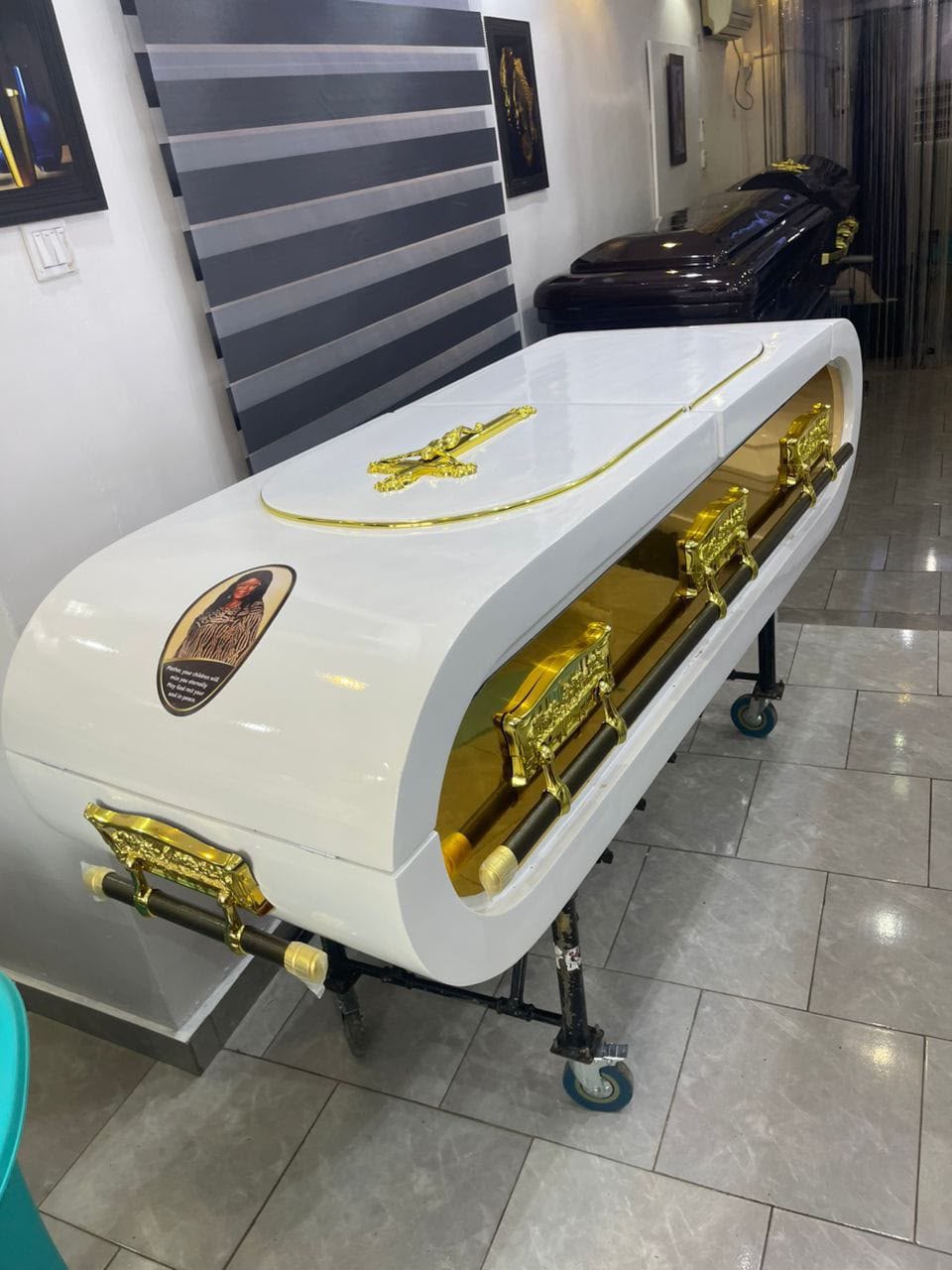 Gloss white capsule-style casket with gold side panels and gold cross ornament on a wheeled bier